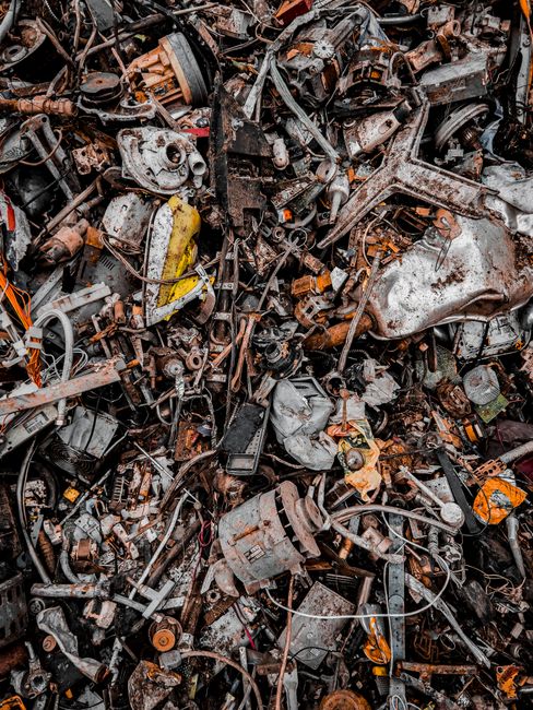A large, densely packed pile of discarded vehicle parts and mechanical debris, including rusted metal engine components, gears, pipes, and various small electronic parts, all tangled together in a chaotic arrangement. The materials exhibit a mix of textures with corroded, weathered surfaces and metallic finishes in shades of brown, orange, silver, and black. The background appears to be an outdoor industrial or scrapyard setting, with the debris filling the entire frame and no clear open space visible. The lighting is diffuse, highlighting the uneven surfaces and rust formations on the metal objects. This scene exemplifies the type of waste managed by private rubbish removal services, such as Rubbish Removal Wandsworth, and reflects the typical clutter associated with vehicle scrap handling and metal recycling operations.
