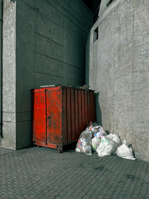 A red metal debris container with visible signs of rust and weathering, positioned against a grey wall on a textured concrete sidewalk. To the right of the container, there are several white plastic garbage bags filled with rubbish, some of which are partially opened, revealing mixed waste inside. The background includes a dark green building wall with horizontal paneling on the left and a light grey, rough-textured concrete wall on the right, with a small rectangular window near the top of the grey wall. The scene appears to be outdoors in an urban setting, likely an alley or side street area, where private waste disposal and rubbish removal services might be employed to manage bulky waste or refuse outside typical collection points. The lighting is natural, suggesting daytime with even illumination across the scene, supporting the context of independent waste handling options like rubbish removal services. Rubbish Removal Wandsworth could handle such waste clearance as part of their specialist rubbish removal offerings, particularly when dealing with non-standard or bulky items outside regular council collections.
