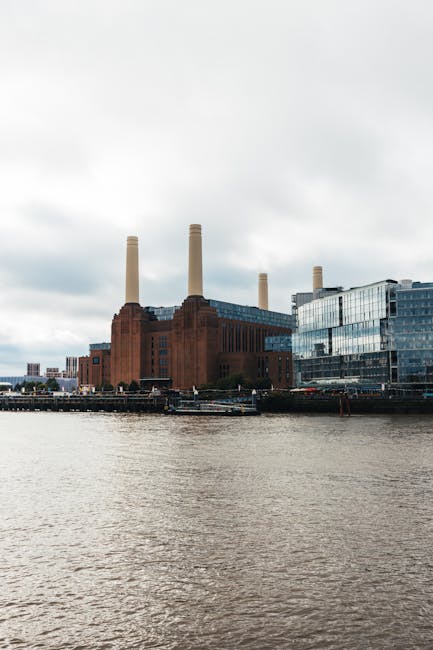 A large, historic power station with five tall, beige-colored chimneys standing vertically against a cloudy sky. The building is made of reddish-brown brick with a modern glass extension on the right side, reflecting the surrounding environment. The power station is situated on the edge of a river, with the water in the foreground displaying gentle ripples. Small boats and vessels are moored along the waterfront near the base of the structure. The scene is captured during daylight, providing clear visibility of the industrial architecture and the calm water. The power station's size and solid construction contrast with the sleek, reflective glass addition, illustrating a blend of old and new architecture often seen in urban areas where modern waste and rubbish removal services might operate for on-site clearance or private disposal solutions near such prominent industrial landmarks. Rubbish Removal Wandsworth offers services that support alternative waste handling with careful consideration of local heritage sites like this power station.