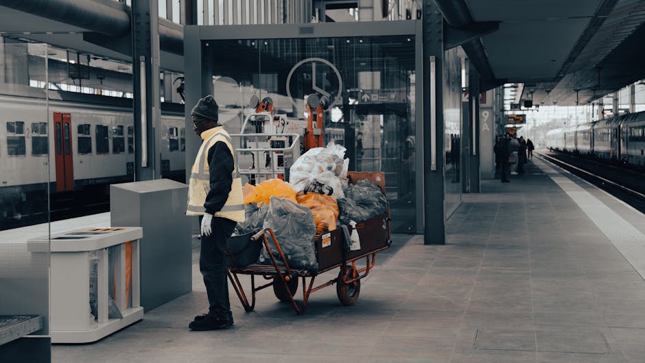 A worker standing on a train station platform next to a large metal trolley loaded with various bags and rubbish, including plastic and fabric trash bags, some orange, black, and transparent, and miscellaneous debris. The worker is wearing a dark jacket, a grey knit beanie, and white gloves, facing slightly to the left with their head turned away. Behind the worker, there is a modern glass-enclosed elevator shaft, with some mechanical equipment visible behind the glass, and a parked train on the left side of the platform. On the right, there are more passengers in the distance waiting or walking along the platform. The surface of the platform is made of grey tiles, and overhead lighting highlights the scene. The environment indicates an urban rail station setting, where private collection of waste may take place, consistent with the context of rubbish removal services by companies such as Rubbish Removal Wandsworth.