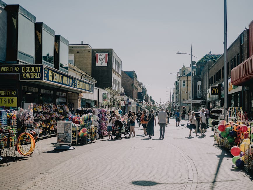 A busy street scene with pedestrians walking along the pavement on a clear day. On the left side, there are outdoor stalls displaying colorful artificial flowers, beach balls, and hula hoops, with merchandise arranged on tables and racks. Behind the stalls, a shop with the sign 'Discount World' is visible, alongside other storefronts, including a McDonald's on the right side. The street is lined with multi-story buildings featuring various commercial signs, some with modern glass facades and others with traditional brickwork. Streetlights and a clear blue sky overhead contribute to the bright atmosphere. The scene captures a typical shopping area, with pedestrians of different ages, some pushing strollers or carrying shopping bags, engaging with the marketplace environment, illustrating an active retail environment frequently associated with independent and alternative waste handling where rubbish might be collected separately from the businesses, in line with private rubbish removal services such as those offered by Rubbish Removal Wandsworth. The overall setting emphasizes urban retail activity and pedestrian use, with no visible waste or litter but implied context of waste management and disposal solutions.
