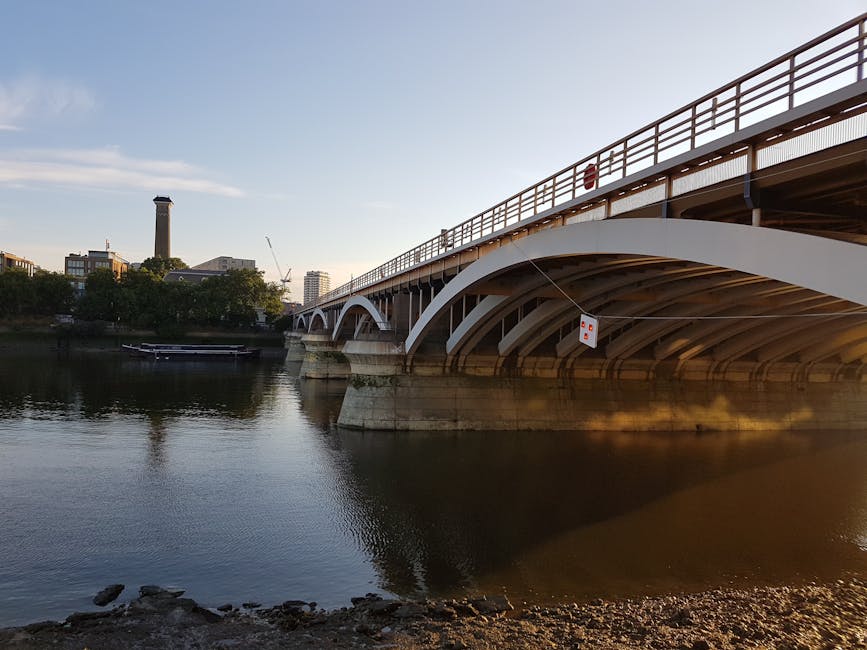 A view of a modern white arch bridge spanning over a calm river during daylight, with the underside of the bridge showing curved supports and a deck with a railing on top. In the background, there are various buildings, including a tall chimney-like structure, and a crane, indicating an urban area. The river's surface reflects the bridge and surrounding structures, while rocks and pebbles are visible along the riverbank in the foreground. The scene is illuminated by natural daylight, casting soft shadows under the bridge. The image appears to relate to alternative waste handling or infrastructure that supports urban development, with the bridge potentially serving as a key piece of local transport or logistics. The scene's clarity and detail focus on the concrete and steel construction of the bridge and the tranquil water beneath, which can be associated with the transportation and disposal context managed by waste removal services such as Rubbish Removal Wandsworth.